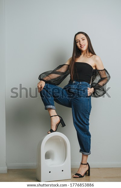 Attractive young woman in fashionable clothes\
standing on a toilet bowl. She\'s wearing moms jeans, heeled\
sandals, net puffed\
sleeves.