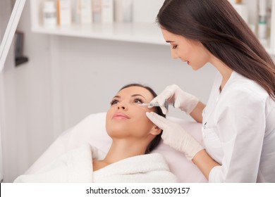 Attractive young woman is getting a collage injection in her face. She is sitting calmly at clinic. The expert beautician is filling female nasolabial wrinkles by hyaluronic acid and smiling