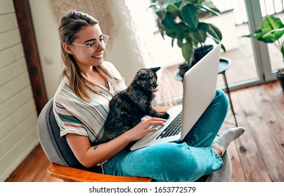 An attractive young woman in glasses is working on a laptop while sitting cross-legged in a comfortable chair at home with a funny assistant cat on her legs.