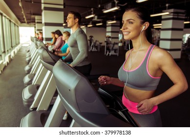 Attractive Young Woman In Headphones Running On A Treadmill In Gym And Smiling