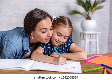 Attractive Young Woman And Her Little Cute Daughter Are Sitting At The Table And Having Fun While Doing Homework Together.