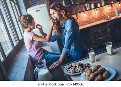 Attractive young woman and her little cute daughter are eating cakes and cookies on kitchen and drinking milk. Having fun together while enjoying freshly baked pastries.