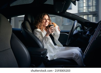 Attractive young woman holding cup of coffee and croissant while sitting on driver seat in electric car with vehicle autopilot system. Cheerful businesswoman having breakfast in electric vehicle.