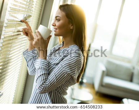 Attractive  young woman holding cup with hot tea or coffee and looking at the sunrise standing near the window in room