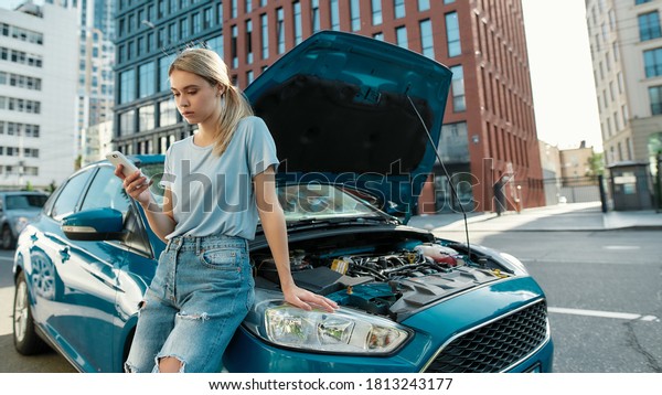 Attractive young\
woman holding phone, calling emergency car service while standing,\
leaning on her broken car with open hood on the city street,\
Selective focus, Web\
Banner