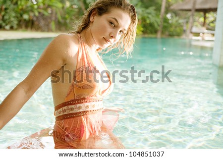 Attractive young woman inside a swimming pool in a tropical garden, wearing a pink dress and smiling.
