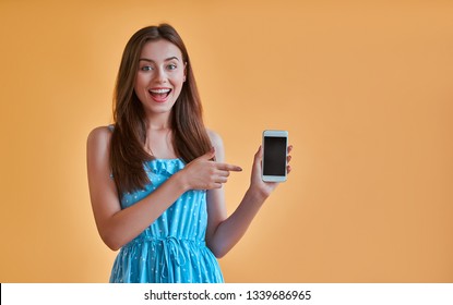 Attractive young woman isolated on orange background. Happy girl in blue dress is posing with smart phone on yellow background.