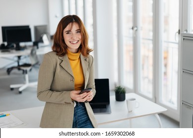 Attractive Young Woman In Jeans, Sweater And Blazer, With Smartphone In Her Hands, Standing Next To The Desk In Office Interior. Looking At Camera And Smiling With Friendly Face. Copy Space