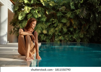Attractive young woman kneeling by the edge of a swimming pool, touching the calm water with her hand