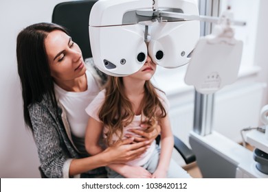 Attractive young woman and little cute girl are checking the eye vision in modern clinic. Mom with daughter in ophthalmology clinic