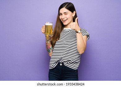 Attractive young woman making a thumbs up and feeling happy while drinking a cold beer. Hispanic woman winking and holding a mug of beer