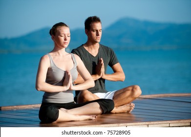 An attractive young woman and man doing yoga on a jetty with the blue ocean and another island behind them - Powered by Shutterstock