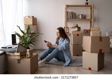 Attractive young woman is moving, sitting among cardboard boxes, using a smartphone and smiling, communicate via smartphone after moving to new flat. - Shutterstock ID 2171273857