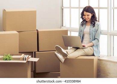 Attractive young woman is moving, sitting among cardboard boxes, using a laptop and smiling