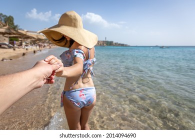 An Attractive Young Woman On The Seashore In A Swimsuit And A Big Hat Walks By The Hand With A Guy.