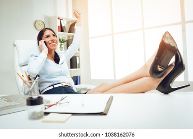 Attractive Young Woman With Open Arms Sitting At The Office With Legs Up On A Desk And Celebrating Success. 