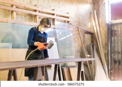 Attractive Young Woman Painting A Table With A Spray Gun While Working In A Woodshop