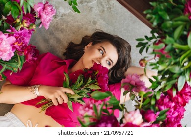 An Attractive Young Woman In A Pink Blouse With Peony Flowers On The Floor. Flower Shop. Training As A Florist. Perfumes For The Body And Home. The Beauty Of Flowers.