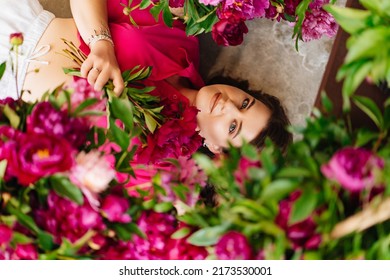 An Attractive Young Woman In A Pink Blouse With Peony Flowers On The Floor. Flower Shop. Training As A Florist. Perfumes For The Body And Home. The Beauty Of Flowers.