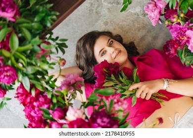 An Attractive Young Woman In A Pink Blouse With Peony Flowers On The Floor. Flower Shop. Training As A Florist. Perfumes For The Body And Home. The Beauty Of Flowers.