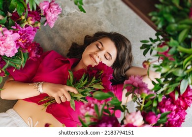 An Attractive Young Woman In A Pink Blouse With Peony Flowers On The Floor. Flower Shop. Training As A Florist. Perfumes For The Body And Home. The Beauty Of Flowers.