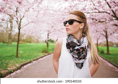 Attractive young woman posing at the spring blossom garden. Pretty caucasian female model standing outdoors at a park wearing sunglasses and scarf looking away.