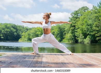 Attractive young woman is practicing yoga, doing Virabhadrasana II pose, standing in Warrior pose near lake in morning.