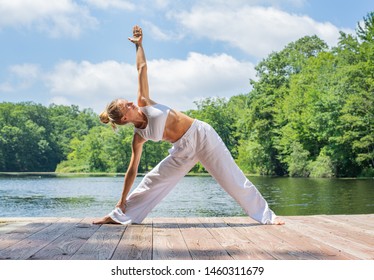Attractive young woman is practicing yoga, doing Utthita Trikonasana pose near lake in morning.