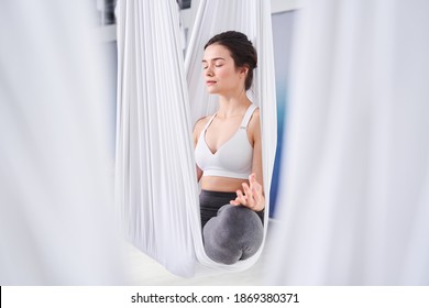 Attractive young woman practicing yoga and doing Ardha Padmasana exercise while sitting on lotus position at the hammock. Woman meditating in half lotus pose with namaste. Yoga concept
