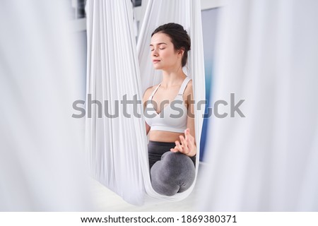 Attractive young woman practicing yoga and doing Ardha Padmasana exercise while sitting on lotus position at the hammock. Woman meditating in half lotus pose with namaste. Yoga concept