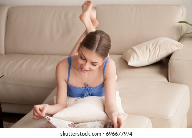 Attractive  Young Woman Reading Beauty Magazine Lying Relaxed On Sofa In Living Room. Pretty Lady Flipping Pages Of Goods Catalog When Resting At Home On Weekend. Female Tourist Relaxing In Hotel Room