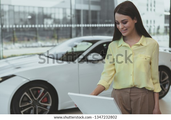 Attractive young woman reading\
info paper about automobile for sale at the dealership salon.\
Beautiful cheerful woman smiling, choosing new car to buy, copy\
space