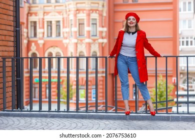 Attractive young woman in a red beret and red coat on a city street against the background of a red building. Woman in jeans, red high-heeled shoes. Fashion portrait. Blurred details
