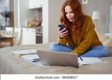 Attractive Young Woman With Red Hair Working From Home - Female Entrepreneur Sitting On Bed With Laptop Computer, Paperwork And Checking Cell Phone From Comfort Of Home