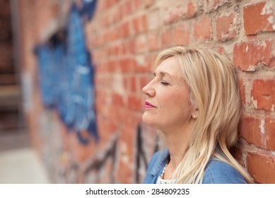 Attractive young woman relaxing leaning back against a brick wall with her eyes closed as she takes a moment for herself, profile view
