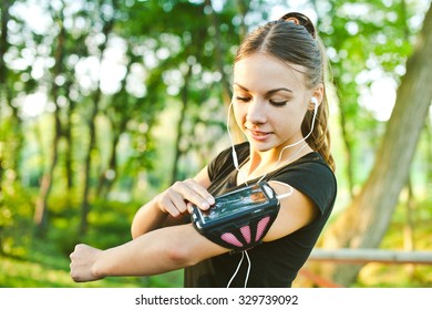 Attractive young woman runner athlete touch her phone in armband during training in evening park with earphones and music at sunset. Pointing, adjusting and looking at gadget display before run - Powered by Shutterstock