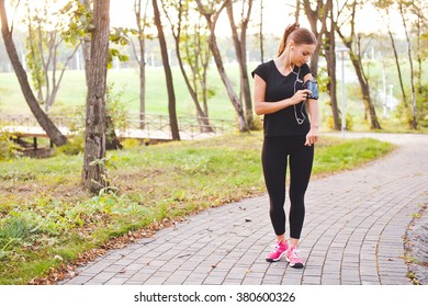 Attractive young woman runner athlete touch her phone in armband during training in evening park on a path with earphones at sunset. Adjust and look at gadget display before run. Full length portrait - Powered by Shutterstock