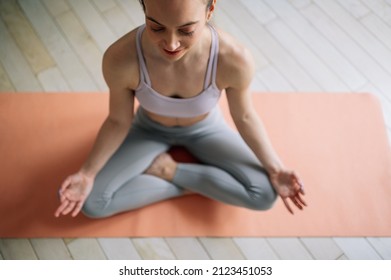 Attractive Young Woman Siting In A Lotus Yoga Position On The Fitness Mat At Home In Living Room. Relaxing State Of Mind, Meditation. No Stress Concept. Top View.