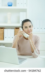 Attractive Young Woman Sitting At Desk In Home Office, Talking On Landline Phone.