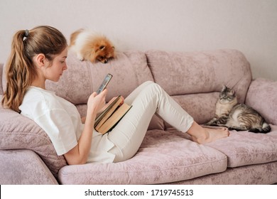 Attractive Young Woman Sitting On The Sofa Reading A Book And Was Distracted By The Phone. The Cat And Dog Are Resting Next To The Owner. Beautiful Living Room