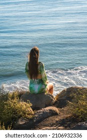 Attractive Young Woman Sitting On A Cliff By The Beach Looking At The Ocean, Malibu, Los Angeles, California