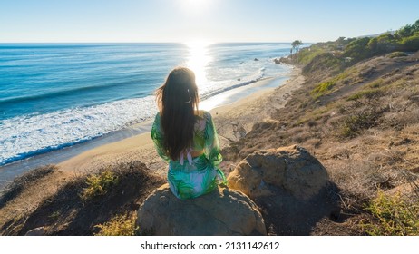 Attractive Young Woman Sitting On A Cliff By The Beach Looking At The Ocean, Malibu, Los Angeles, California