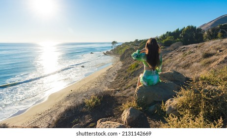 Attractive Young Woman Sitting On A Cliff By The Beach Looking At The Ocean, Malibu, Los Angeles, California