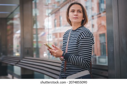 Attractive young woman with smartphone standing at modern bus stop in city