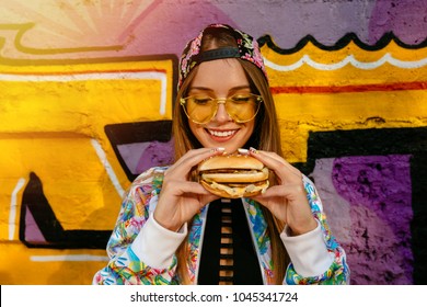 Attractive young woman, smiling cheerfully, holds tasty burger in two hands. Dressed in colorful jacket and cap, in sunglasses. Outdoors, near the wall with graffiti. - Powered by Shutterstock