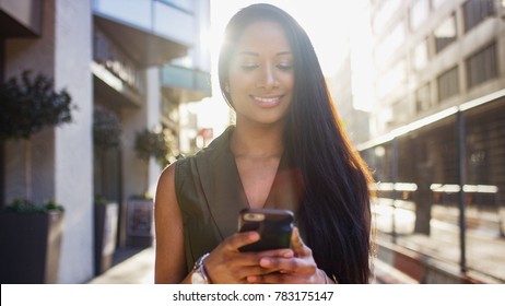Attractive young woman smiling as she uses her phone walking down the street in the city