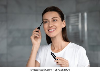 Attractive Young Woman Standing In Bathroom, Looking In Mirror, Applying Black Brown Mascara On Lashes In Morning. Smiling Pretty Brunette Lady Enjoying Doing Daily Nude Makeup Routine At Home.