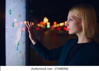 Attractive young woman standing front big digital screen with basic city street map data, female touching sensitive display of modern smart city bus stop for check her location into the night