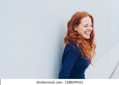 Attractive young woman standing giggling or laughing at something she finds very funny while leaning against a white exterior wall with copy space