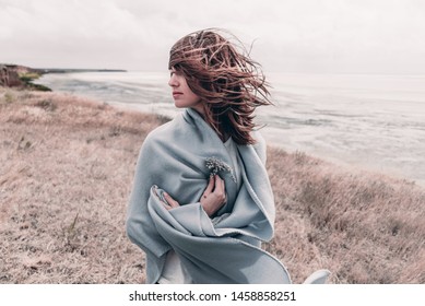 Attractive young woman standing on a windy cold beach wrapped in warm blanket with flower in her hand. - Powered by Shutterstock